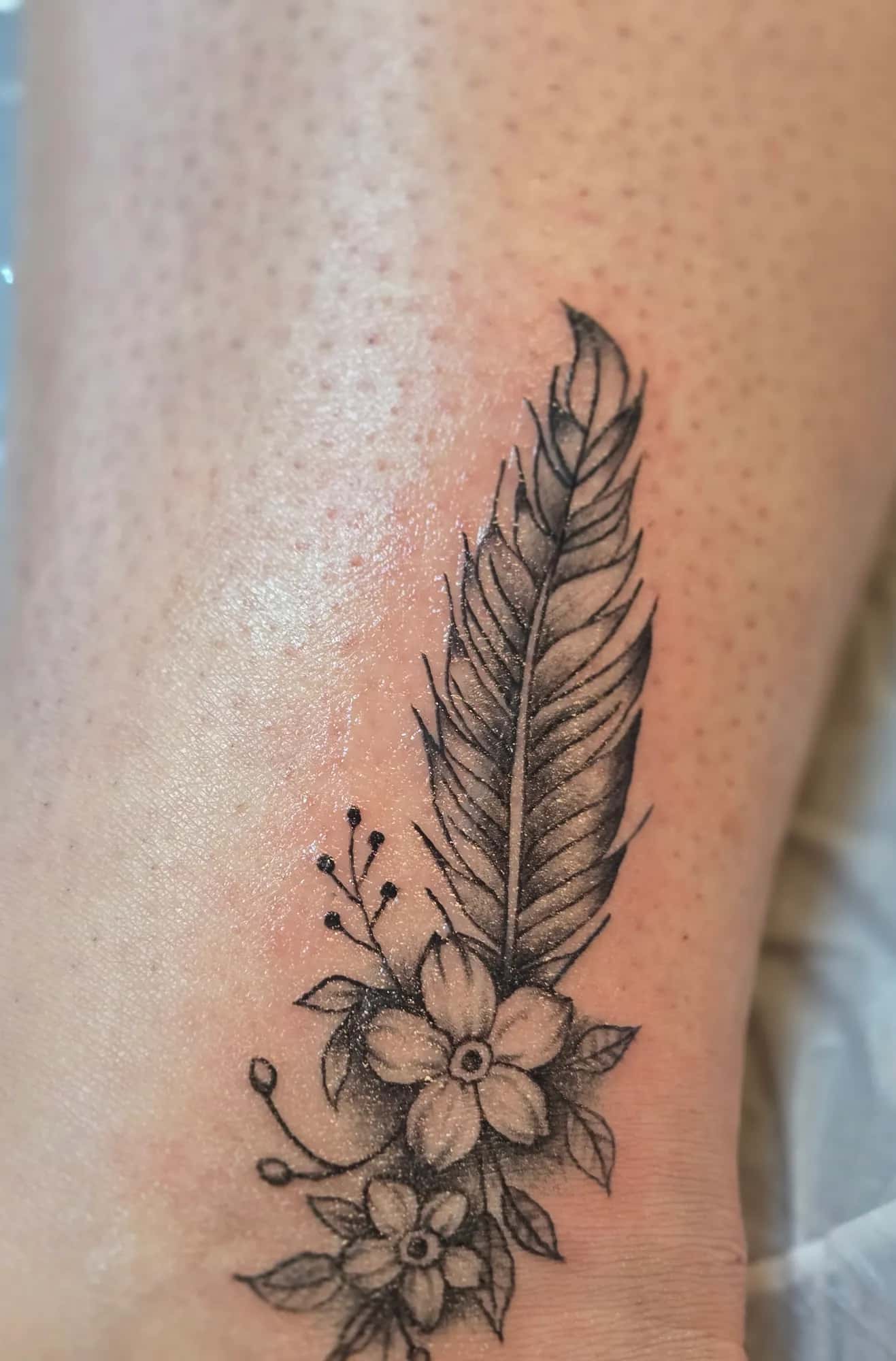 Fineline veer-tattoo met bloemen op de enkel, gezet bij GlowLine in Wilp